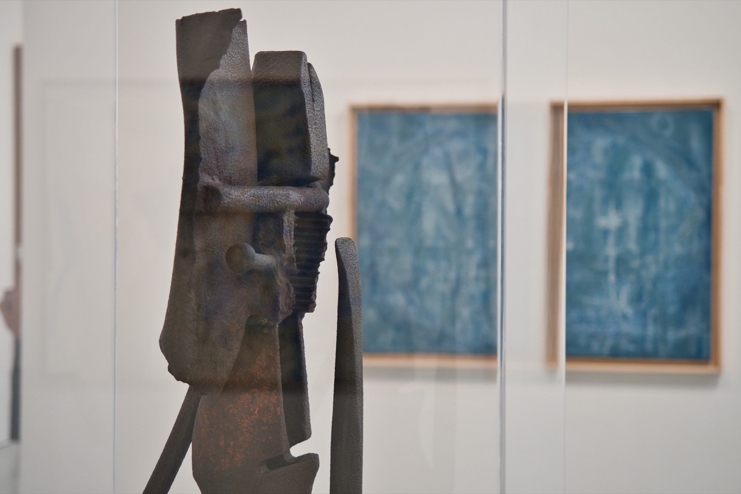 Une sculpture métallique sombre devant des tableaux abstraits bleus.