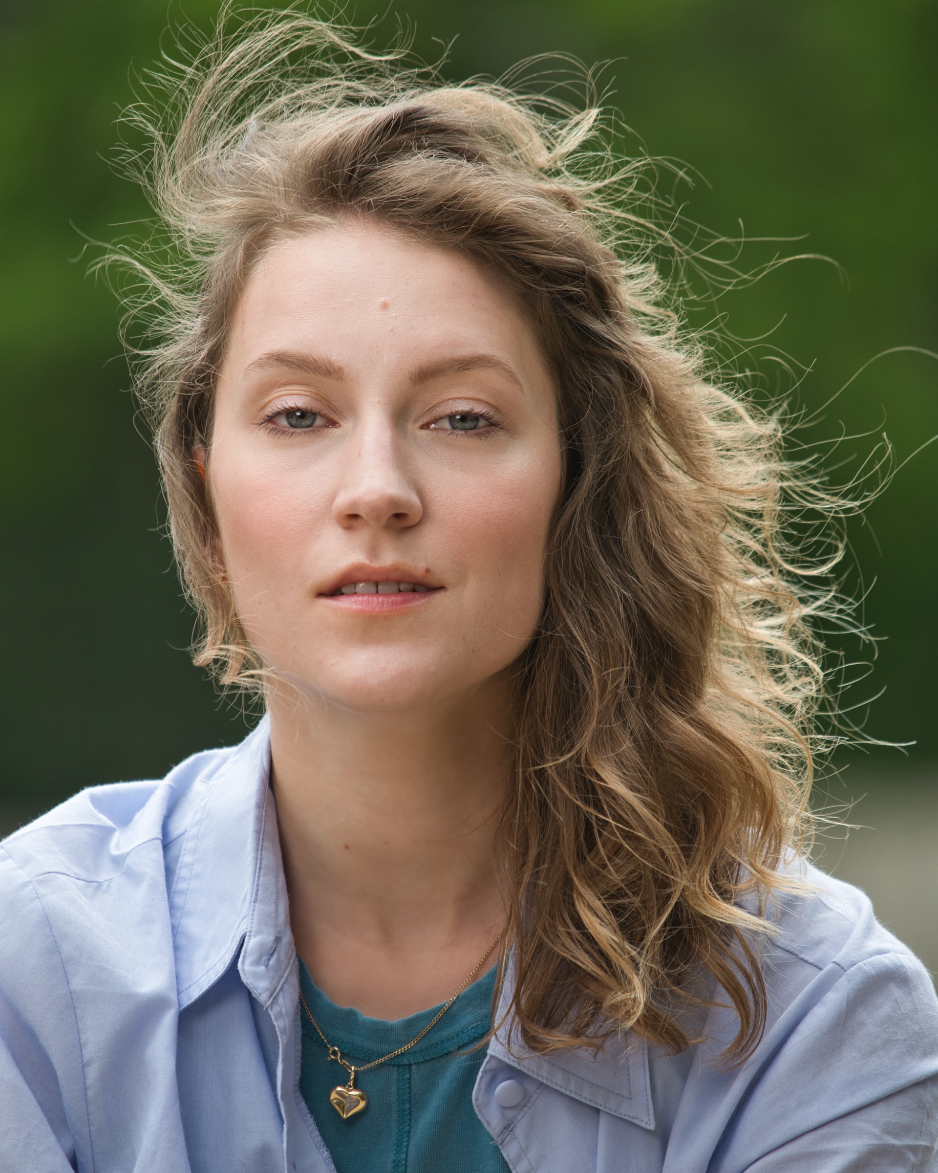 Femme aux cheveux ondulés, regard franc, avec un sourire léger, arrière-plan flou verdoyant.