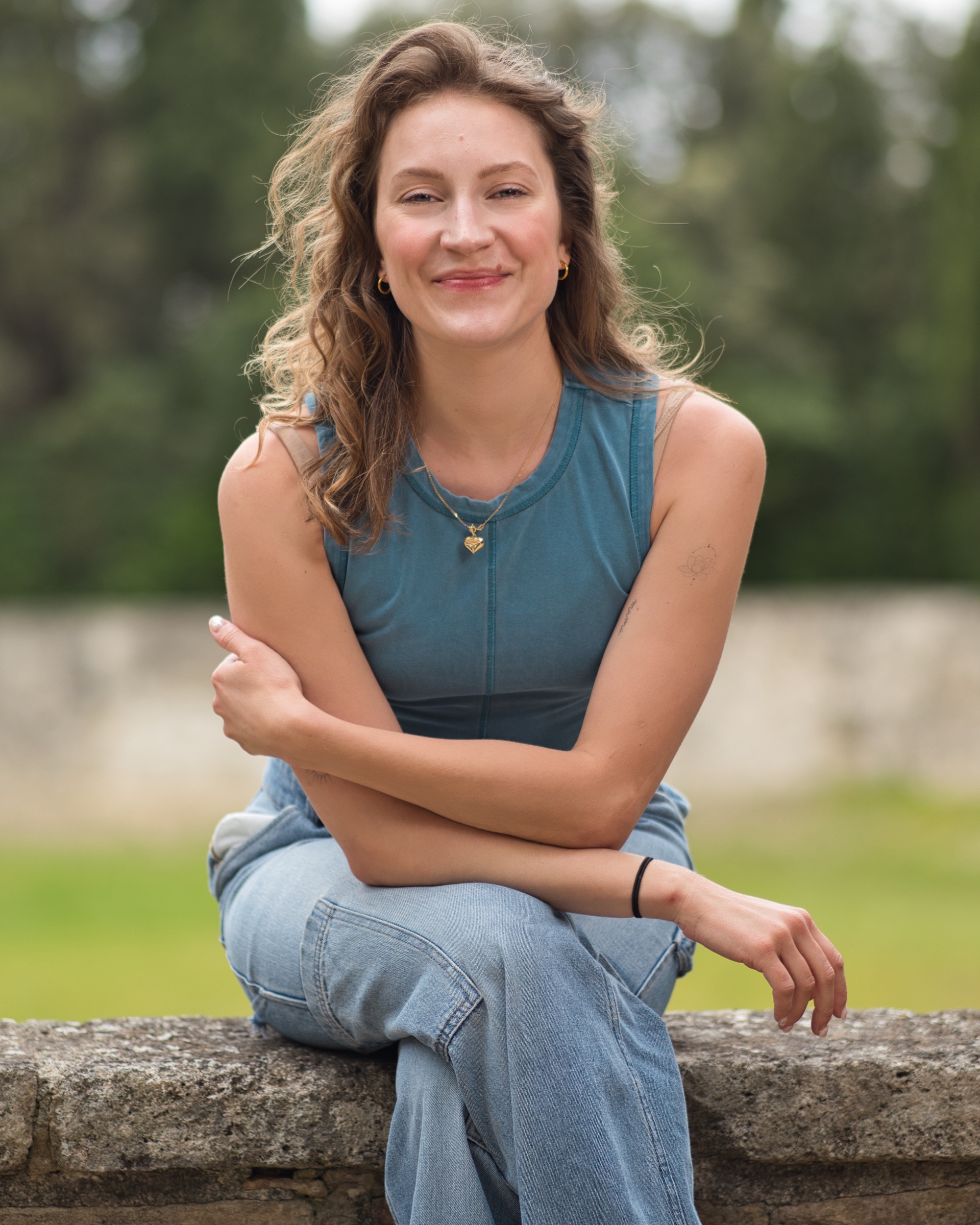 Femme souriante, cheveux ondulés, vêtue d'un top bleu et jeans, assise sur un mur.