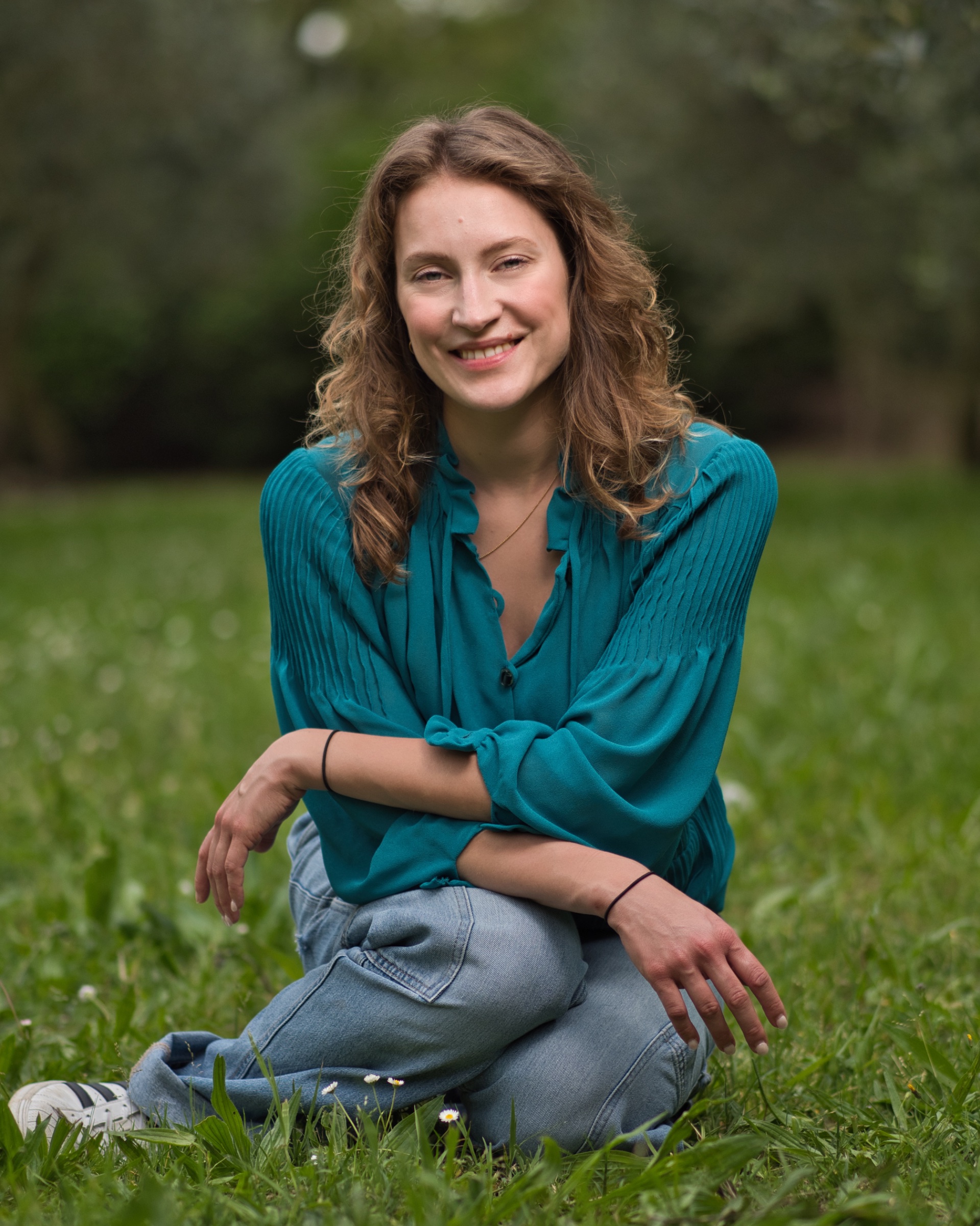 Femme souriante assise dans l'herbe, portant un haut turquoise et un jean.