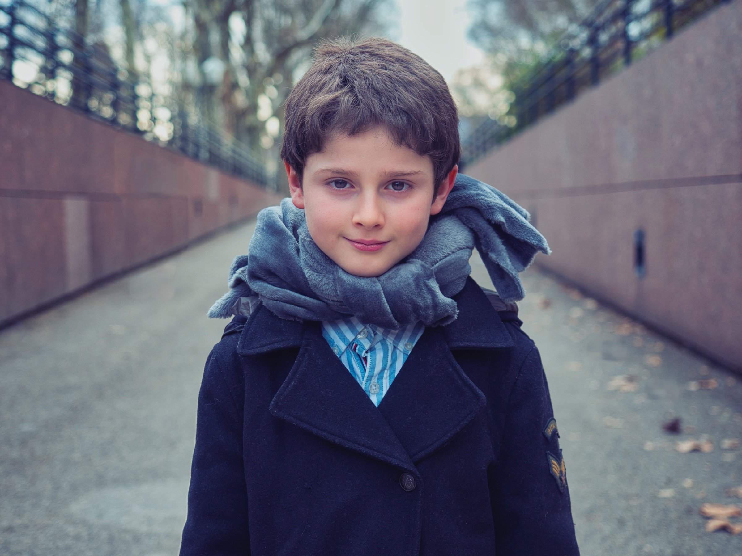 Un garçon souriant en manteau noir et écharpe grise, sur un chemin silencieux.