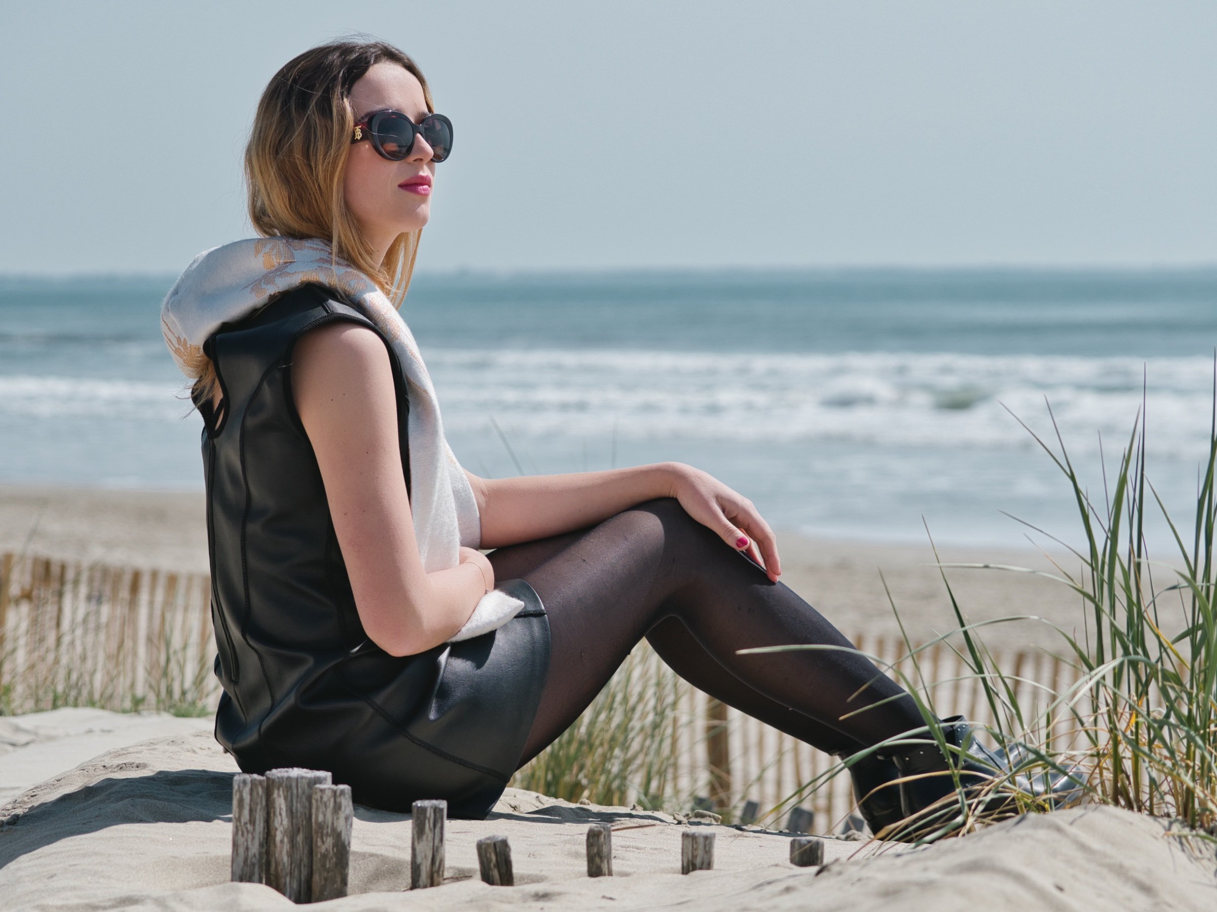 Femme élégante en robe noire, assise sur la plage avec une vue sur la mer.