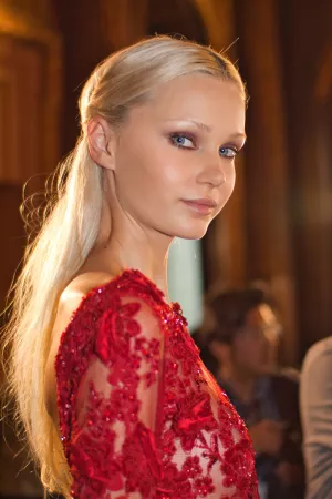 Femme blonde en robe rouge ornée, avec un regard élégant, en arrière-plan flou.
