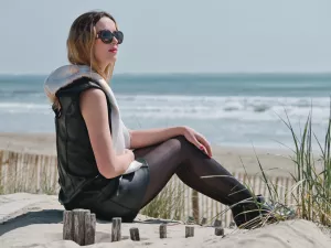 Femme chic en cuir, relaxant sur la plage, lunettes de soleil, mer en arrière-plan.