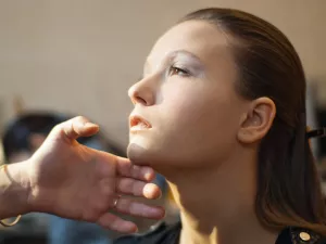 Femme avec un maquillage subtil, la tête légèrement inclinée, main touchant son menton.