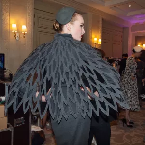 Femme de dos, vêtue d'une cape grise à motifs en plumes stylisées. Ambiance de défilé.