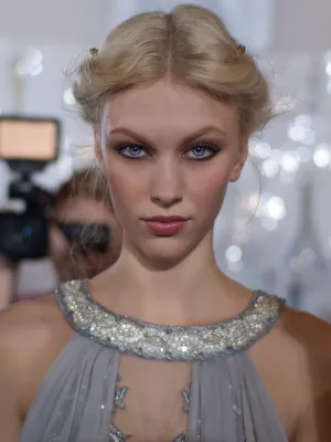 Femme au regard intense, cheveux blonds, robe argentée ornée, fond flou.