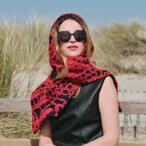 Femme au regard confiant, lunettes noires, écharpe rouge, devant des herbes et un ciel bleu.