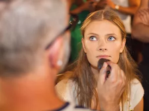 Femme aux longs cheveux blonds se faisant maquiller, visage concentré et lumineux.