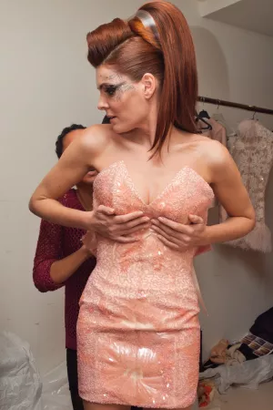 Une femme en robe rose scintillante, avec une coiffure volumineuse, s'apprête pour un défilé.
