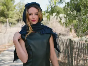 Femme au sourire, portant un manteau en cuir et un châle sombre, sur un chemin bordé d'arbres.