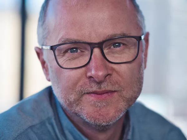 Homme souriant avec des lunettes et une barbe, en arrière-plan flou.